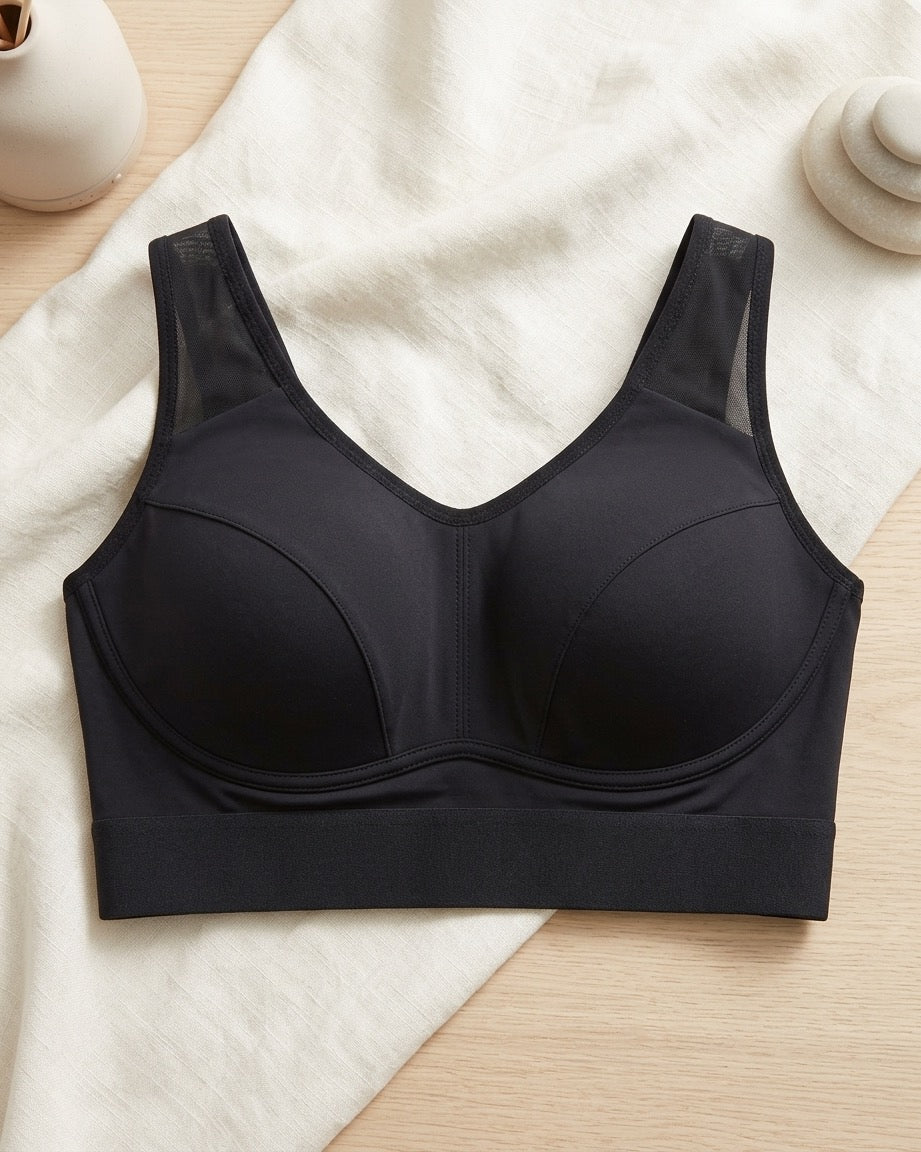A black Mesh Front Leisure Bra, laid flat on a cream fabric surface with a light wood background and small decorative props.